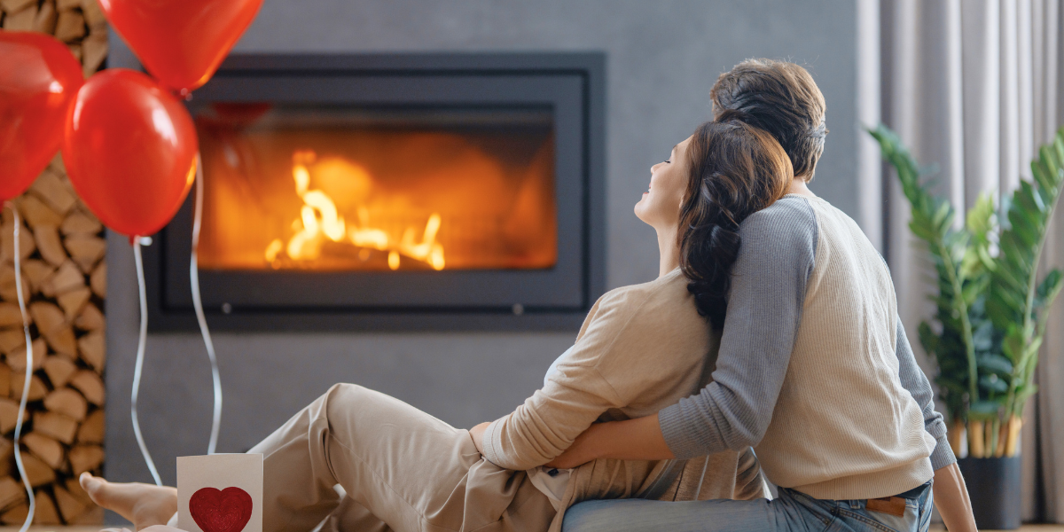 Couple cuddling by a fireplace next to red balloons. 