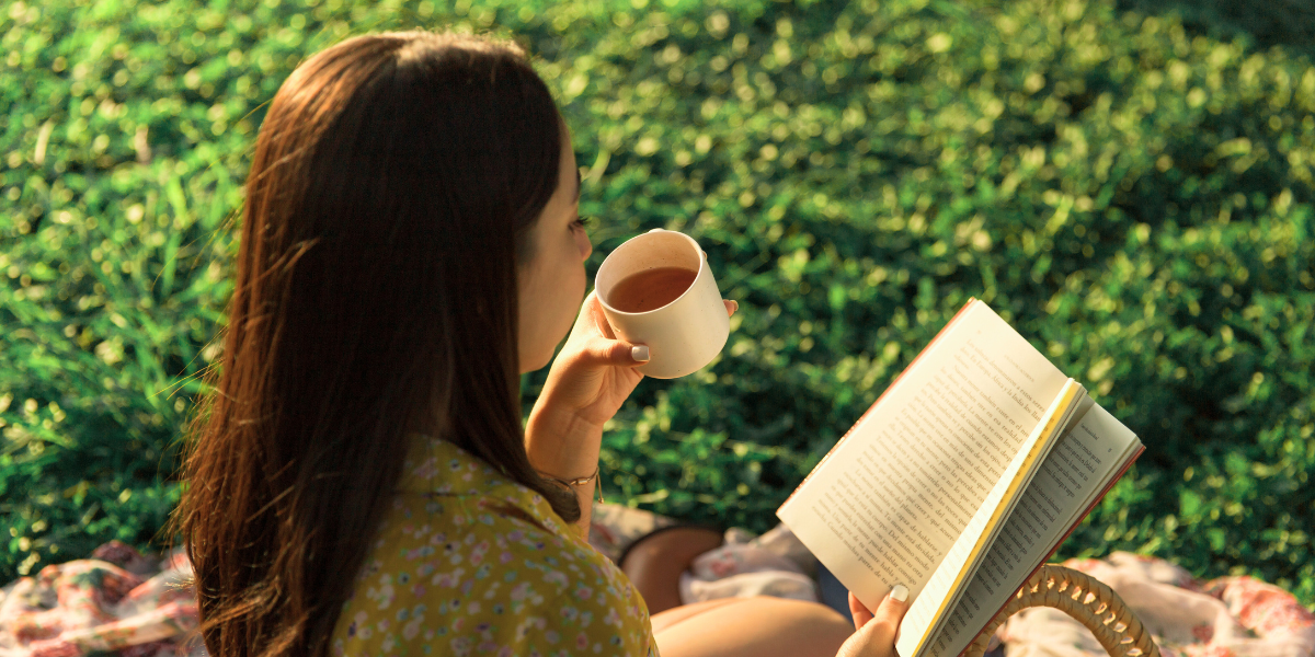 A person enjoys a cup of coffee while reading a book on the grass outside.