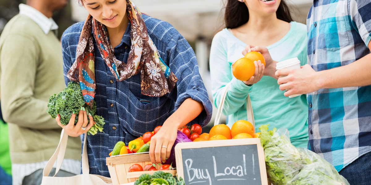 Person sorting through a crate of fresh produce next to a sign that reads "Buy Local!"